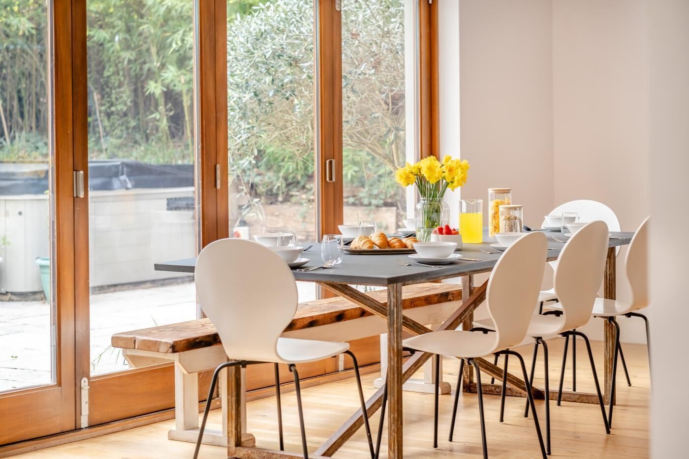 A dining area with a table set for four, chairs, a bench, and a view through large windows.