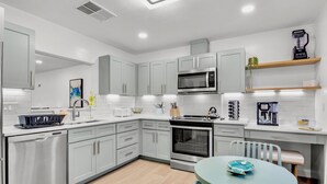 A modern kitchen with stainless steel appliances, white cabinetry, and a dining area with a round table. - Our House at the Beach C-113 (Siesta Key)
