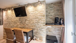Ein Zimmer mit einer Steinwand, einem Flachbildfernseher, Korbsesseln, einem Holztisch, einer Kaffeemaschine und einem Kühlschrank.