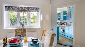 A dining area with a table set for breakfast, a window with a view of greenery, and a kitchen visible through an open door.