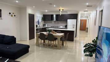 A modern living room with a dark sofa, a dining area with a wooden table and chairs, a kitchen with dark cabinets and a white fridge, and a hallway leading to a door.