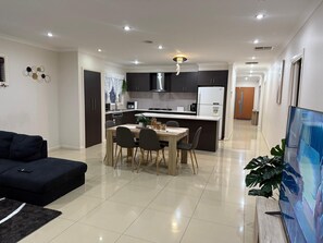Una sala moderna con un sofá oscuro, un comedor con una mesa y sillas de madera, una cocina con armarios oscuros y un refrigerador blanco, y un pasillo que conduce a una puerta.