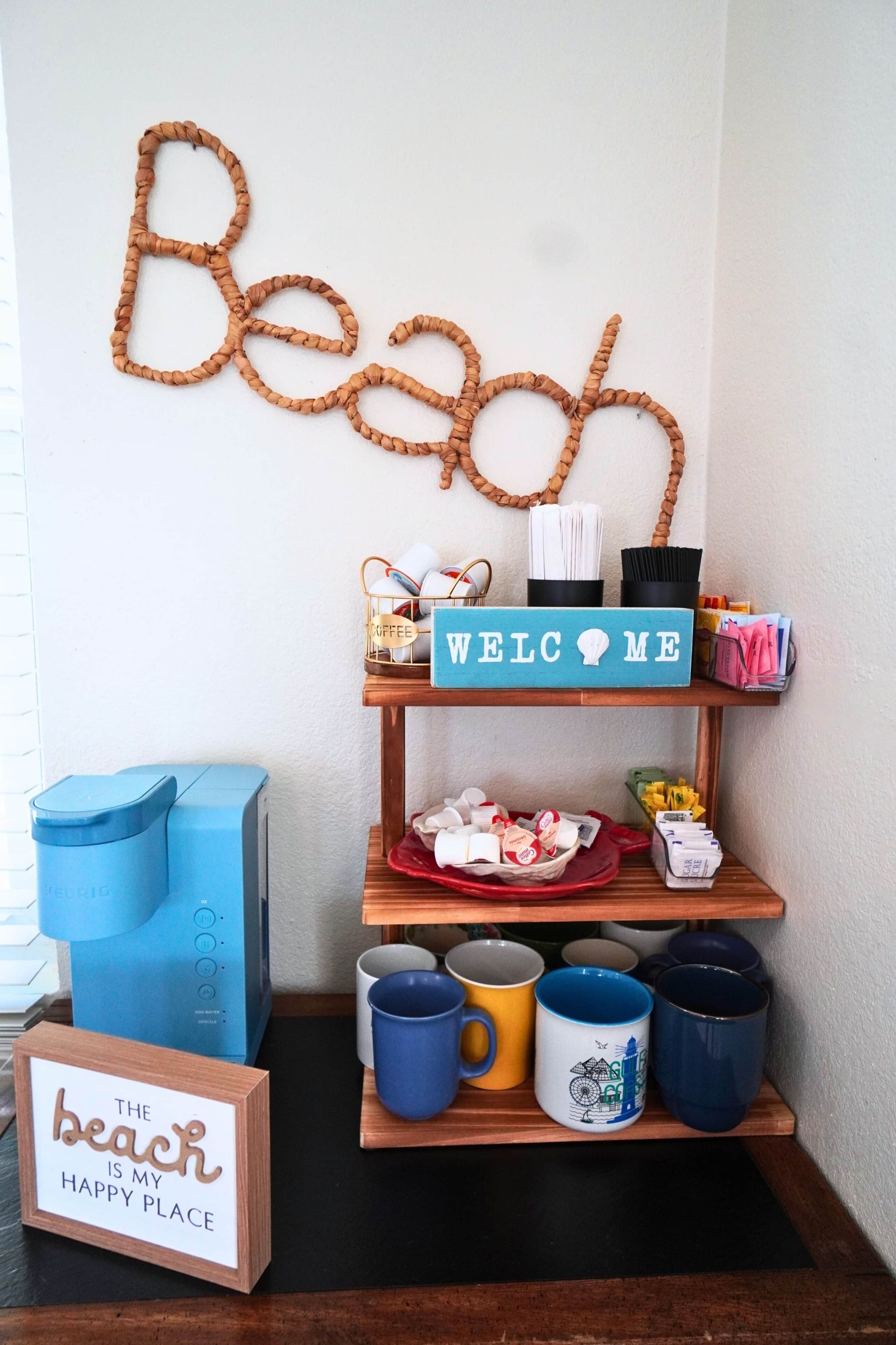 Et værelse med en vægdekoration, der staver 'Beach', et 'Welcome Me'-skilt, en kaffemaskine, krus og et skilt med teksten 'Stranden er mit lykkelige sted'.