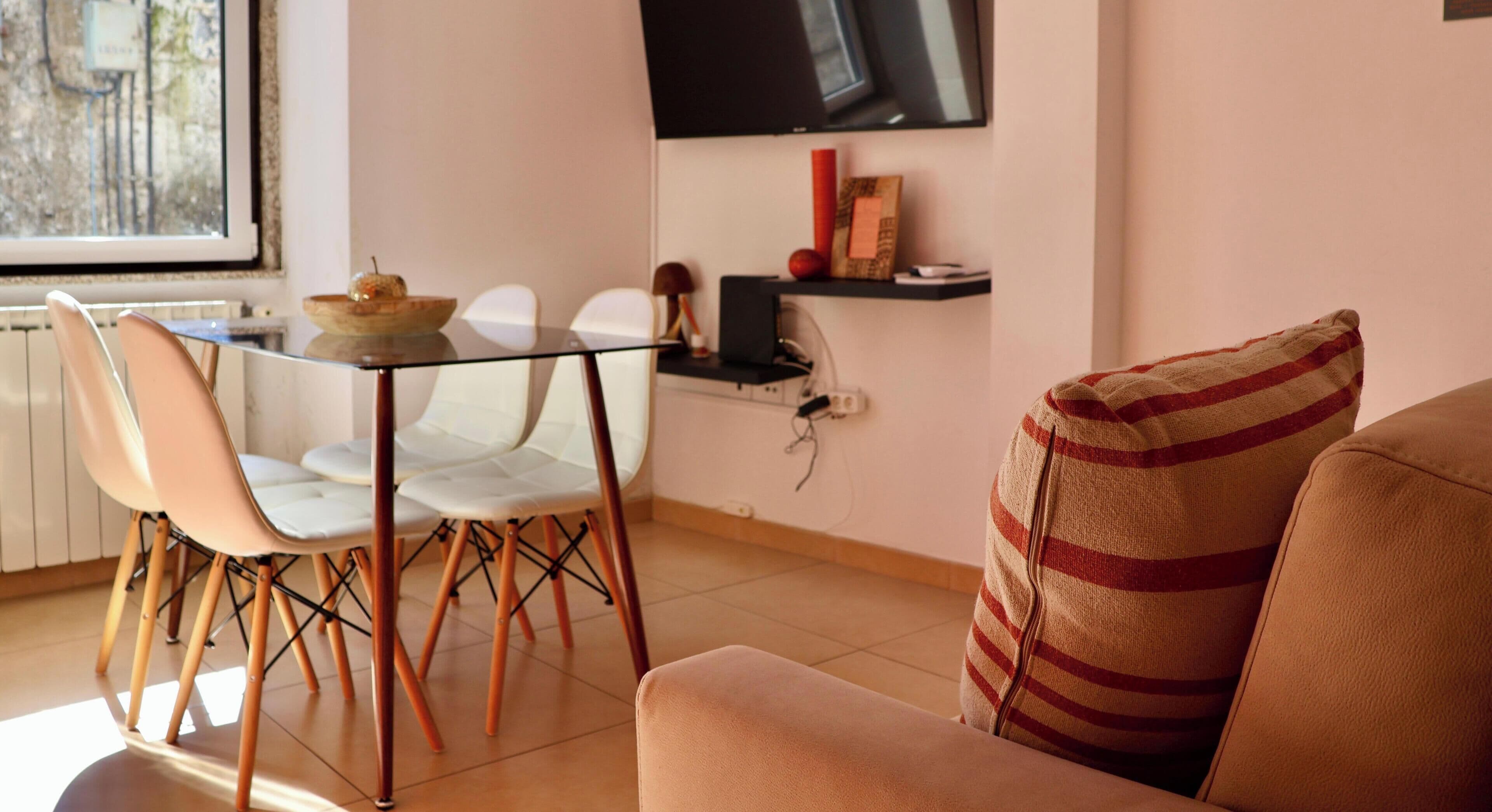 Ein Essbereich mit Glastisch und Stühlen, einem an der Wand montierten Fernseher und einem Sofa mit gestreifter Sitzkissen.