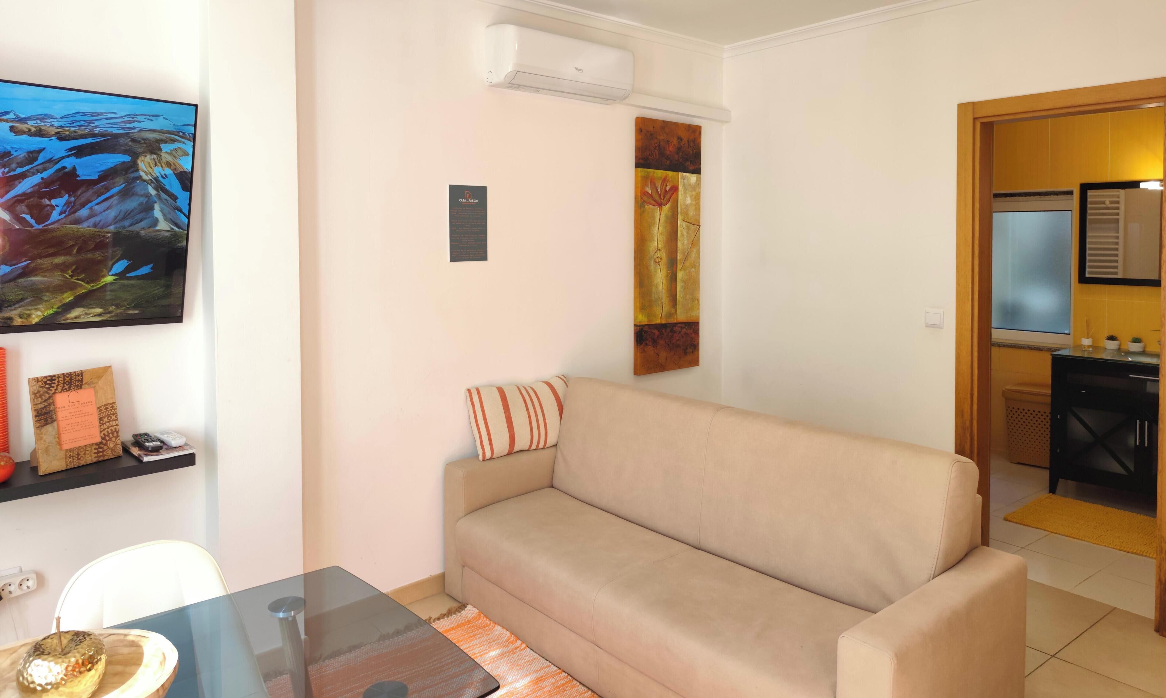 Una sala de estar con un sofá beige, una mesa de centro de vidrio, un cuadro en la pared y una cocina visible a través de un arco abierto.