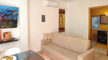 Una sala de estar con un sofá beige, una mesa de centro de vidrio, un cuadro en la pared y una cocina visible a través de un arco abierto.