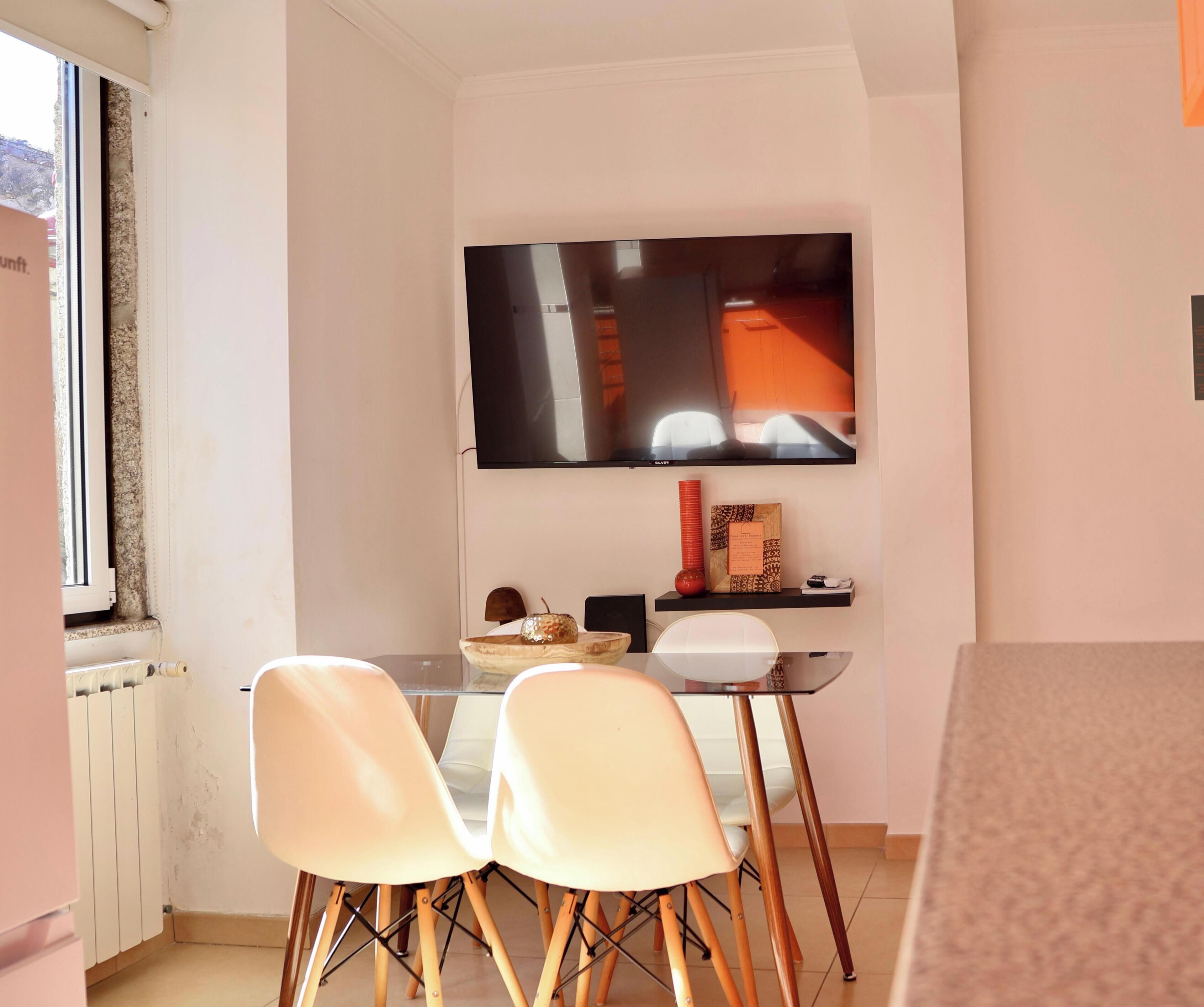 A compact living space with a dining area featuring a glass table and two chairs, a mounted flat-screen TV, and a small shelf with decorative items.