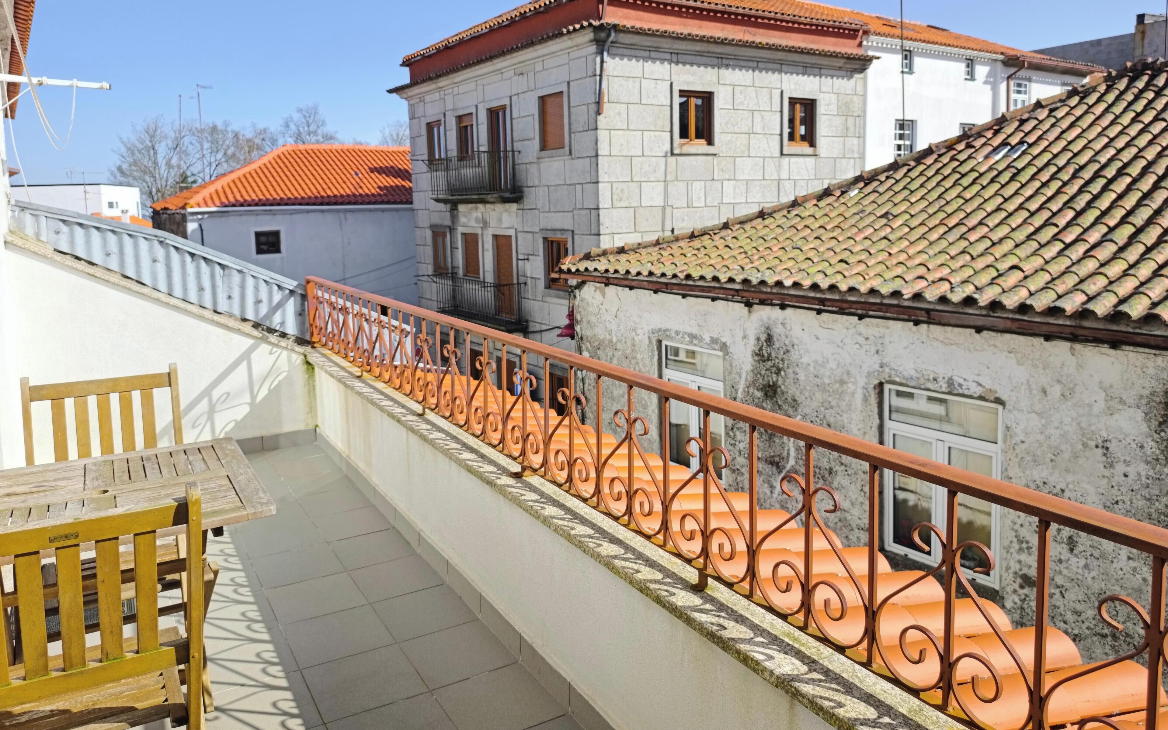Varanda com guarda-corpo em ferro forjado, um banco de madeira e vista para edifícios vizinhos com telhados de telha vermelha.