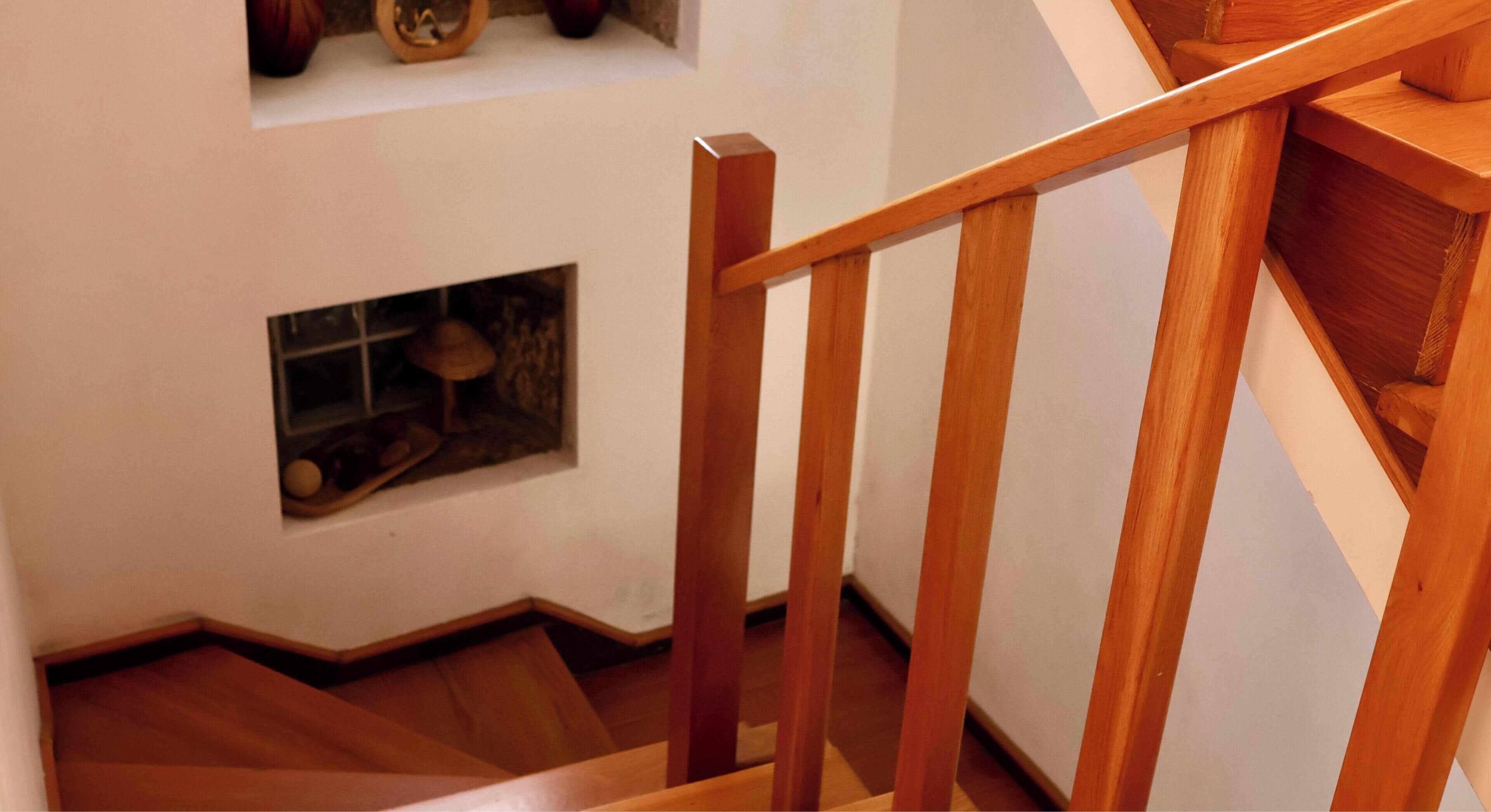Uma escada de madeira com corrimão, que leva a um patamar com uma pequena janela exibindo um objeto decorativo.