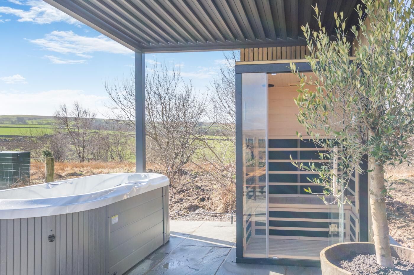 A modern outdoor hot tub area with a wooden deck, a glass door leading to a covered space, and a view of the countryside.