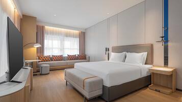 A modern hotel room with a large bed, a desk, a chair, a sofa with patterned cushions, and a television.