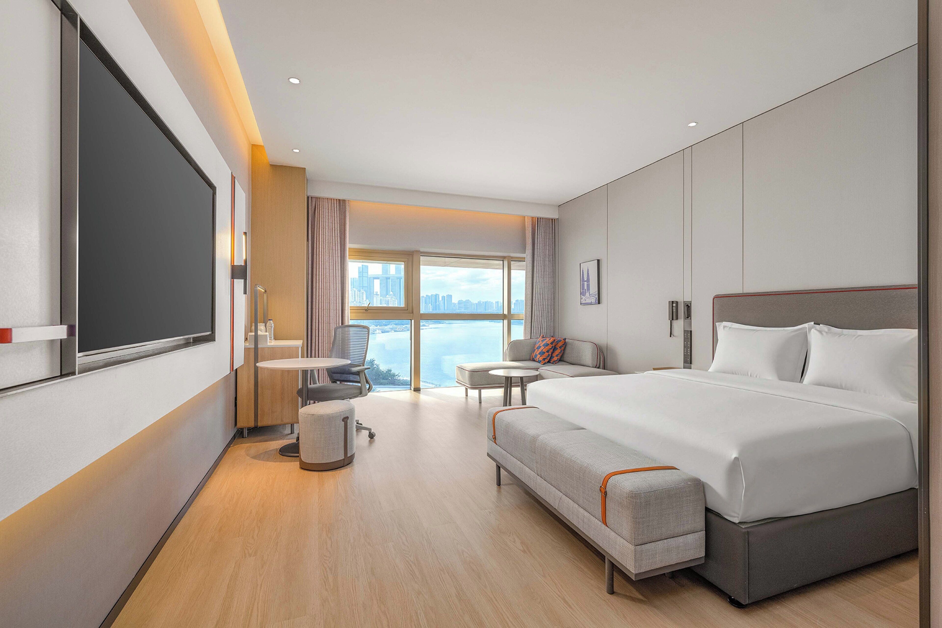A modern hotel room with a large bed, a desk, a chair, and a view of the city.