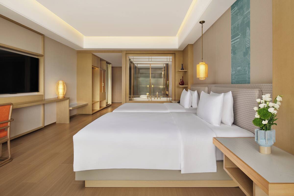 A modern hotel room with a large bed, a desk, a chair, a TV, and a view of the cityscape.