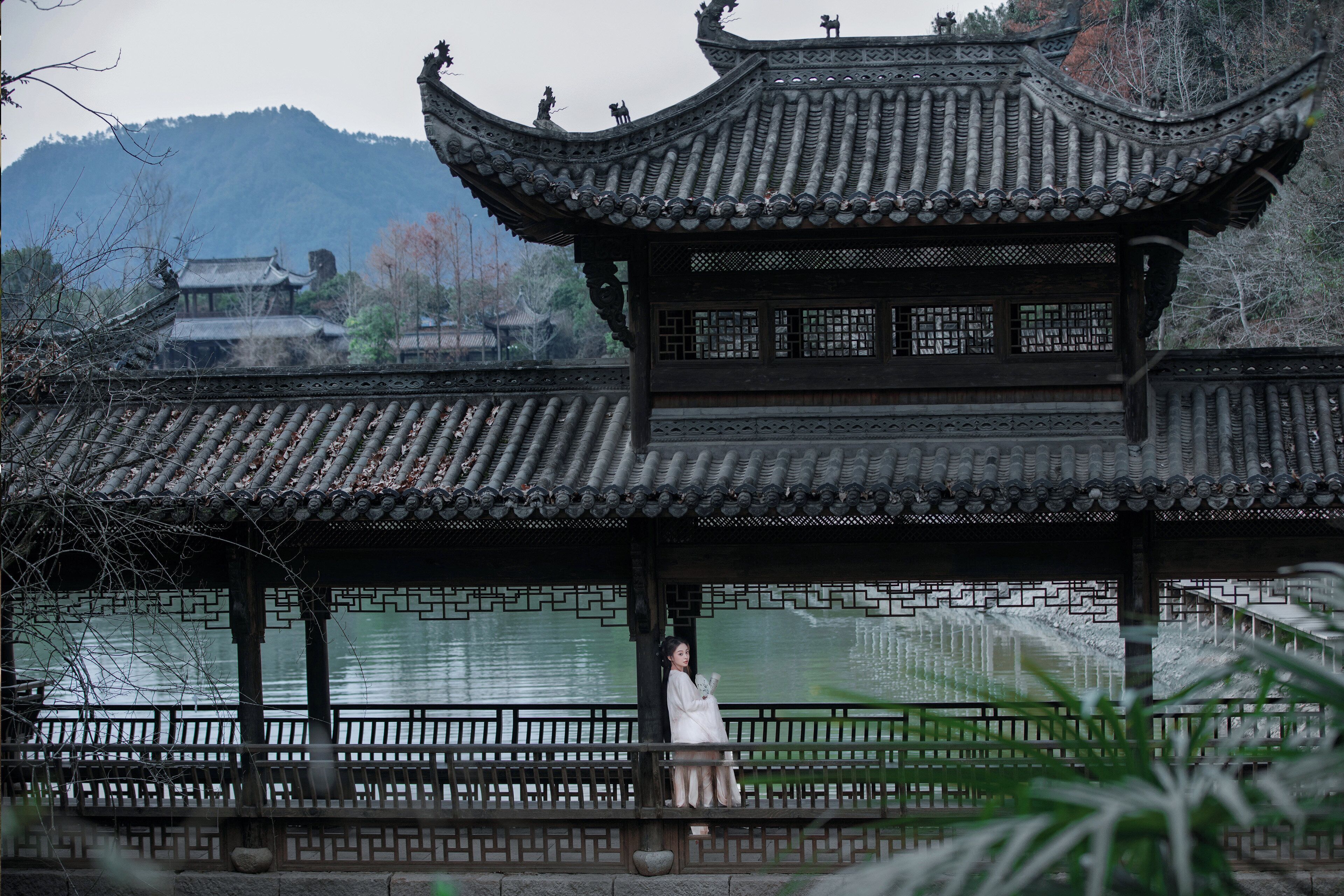 A traditional Chinese pavilion with intricate roof design, a person seated on a bench, and a calm body of water in the background.