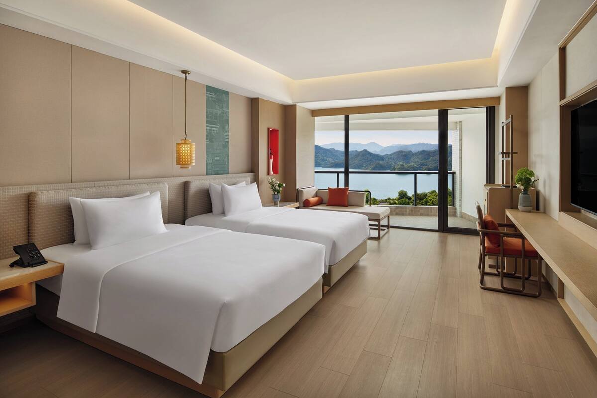A hotel room with two beds, a desk, a chair, a TV, and a large window with a mountain view.