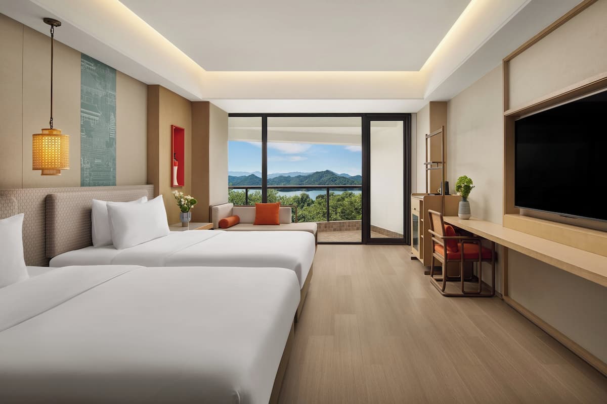 A hotel room with two beds, a TV, a desk, and a balcony with a mountain view.