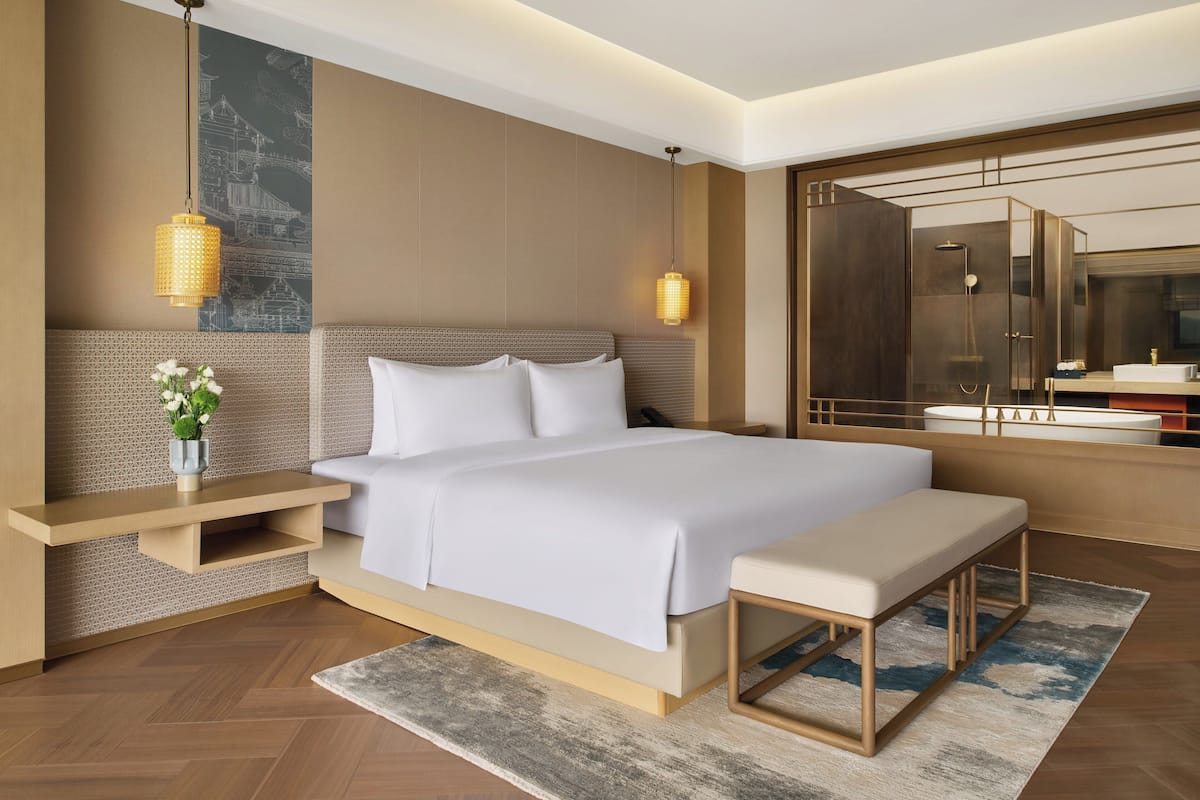 A modern hotel room with a large bed, bedside tables, a bench, a mirror, and a shower area.
