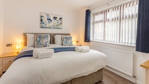 Ein ordentlich bezogenes Bett mit Kissen, eine Nachttischlampe, ein Wandbild, ein Fenster mit Jalousien und eine Heizung.