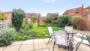 Ein Garten mit einer Pflasterterrasse, einem Tisch und Stühlen, umgeben von einer Ziegelmauer und verschiedenen Pflanzen.