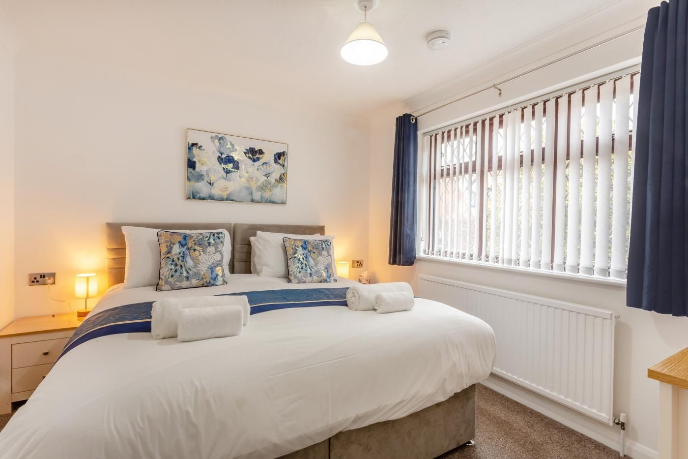 A neatly made bed with pillows, a bedside lamp, a wall-mounted artwork, a window with blinds, and a radiator.