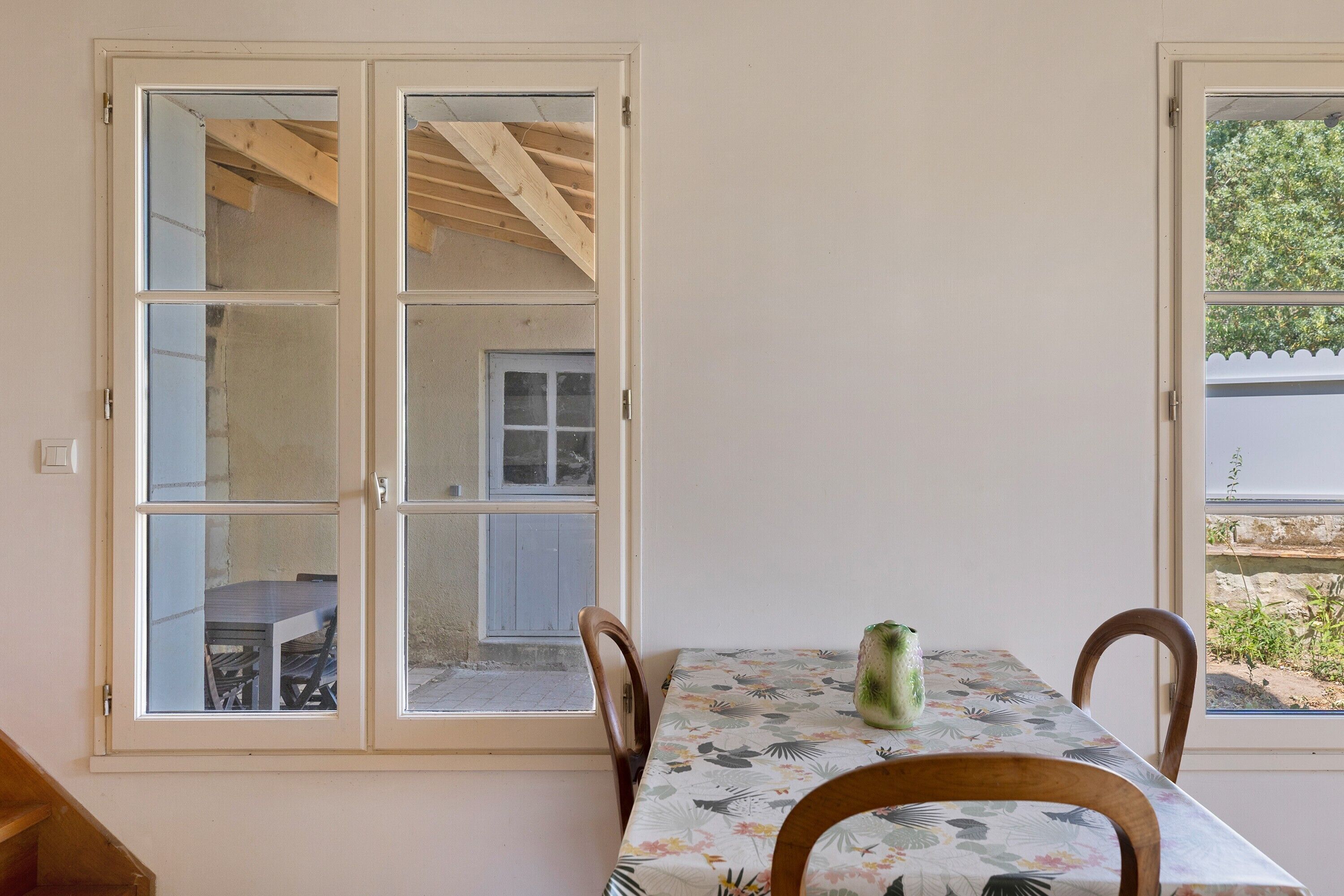 Un coin repas avec une table et des chaises, une fenêtre donnant sur l’extérieur et une pièce dont on aperçoit une partie, avec des poutres en bois apparentes.