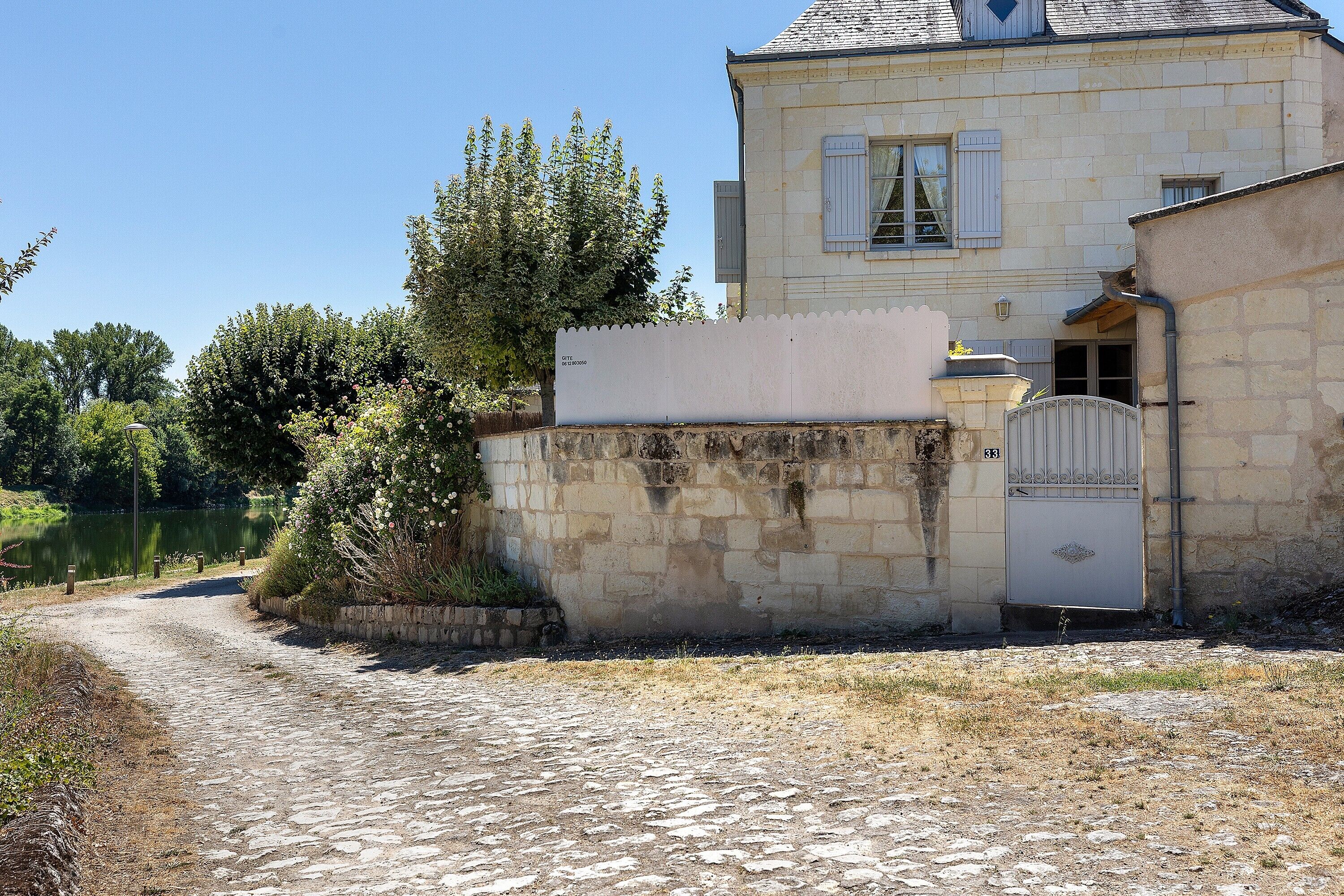 A stone pathway leading to a building with a gated entrance, a small garden, and a body of water in the background.