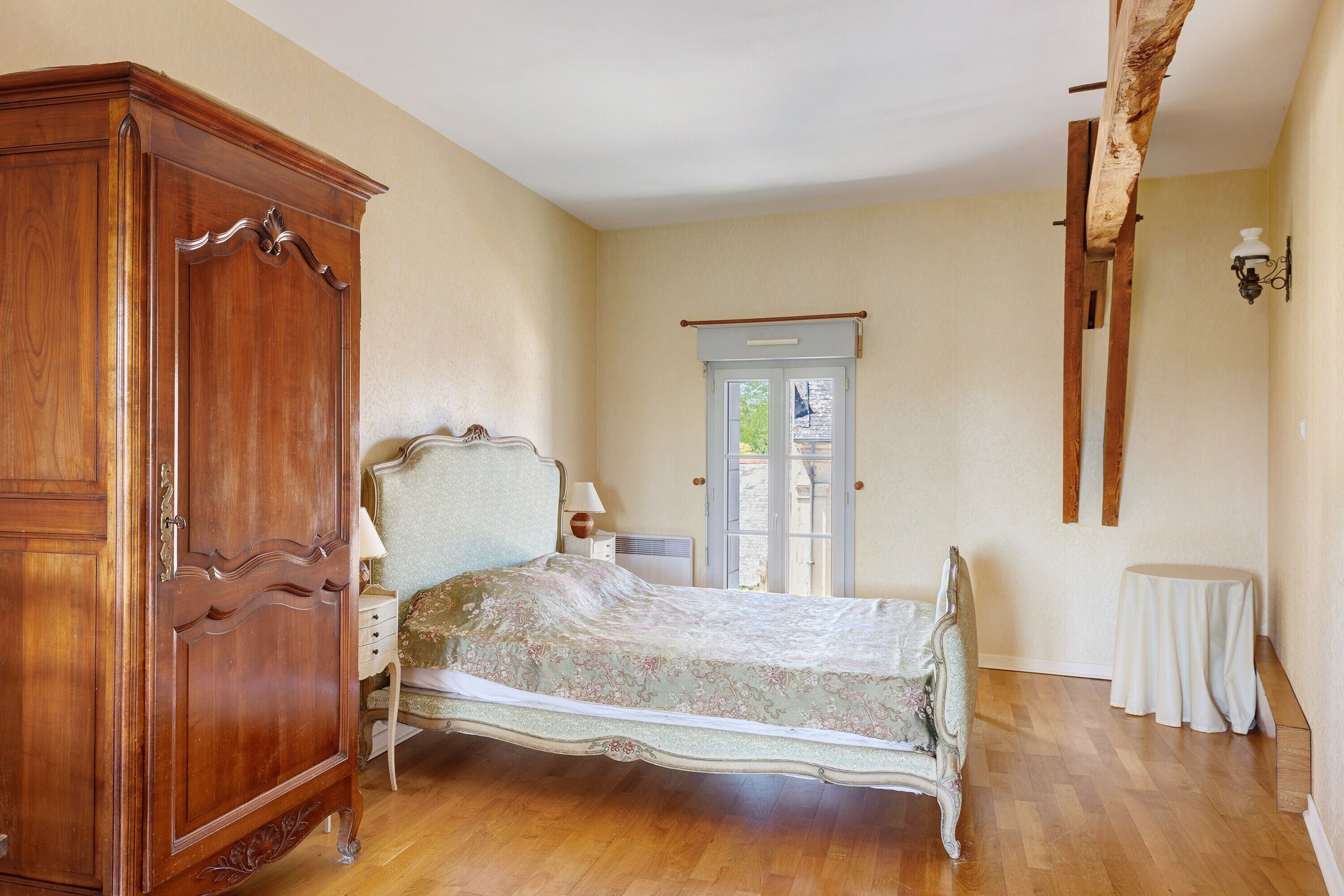 A bedroom with a wooden wardrobe, a bed with a floral bedspread, a small bedside table, a nightstand, a window with a view, and a wall-mounted light fixture.