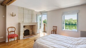 A bedroom with a fireplace, a bed, a chair, and a view of greenery through the window.