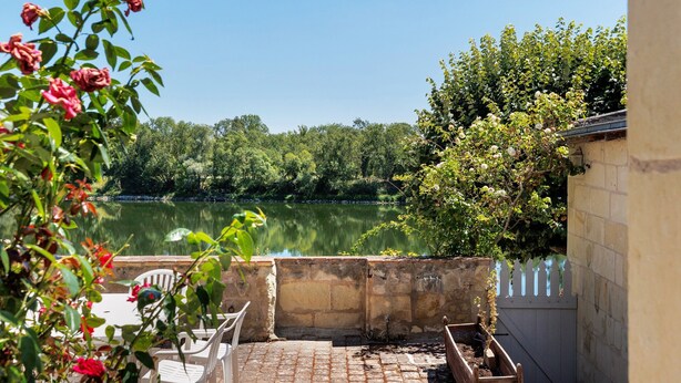 A patio with a view of a calm lake, surrounded by greenery and a stone wall.