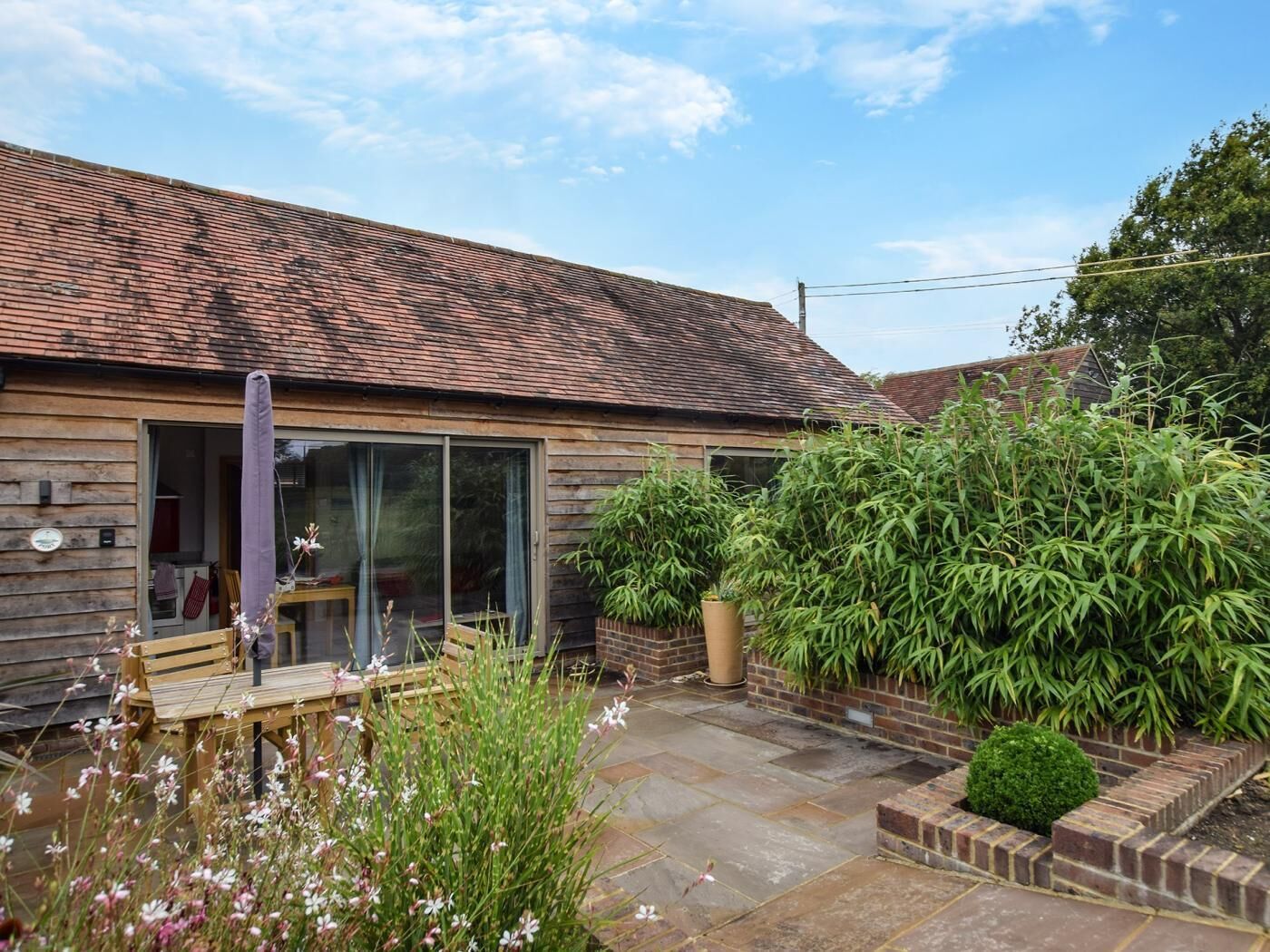 A wooden house with a tiled roof, a glass door, a patio with a table and chairs, and various plants.