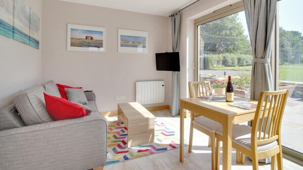 A modern living room with a sofa, a dining table with chairs, a TV mounted on the wall, and a window with curtains. - 1 Bed in Tenterden (oc-79130) (Tenterden)
