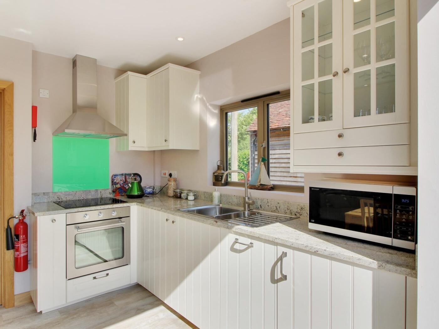 Cocina con armarios blancos, extractor de acero inoxidable, microondas y ventana con vista a zonas verdes.