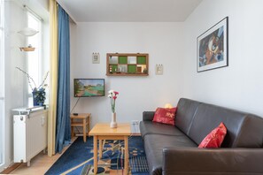 Ein Wohnzimmer mit Sofa, einem kleinen Tisch, einer Heizung, einem Fenster mit Vorhängen, einem Bild und einem Spiegel an der Wand.