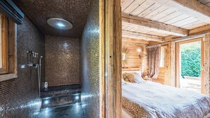 A wooden cabin interior with a shower, a bed with a leopard print bedspread, and a view of greenery outside. - Chalet Zibeline (Megève)