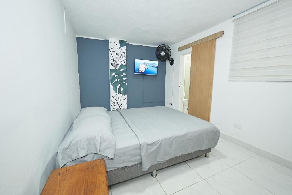 Un dormitorio con cama, televisor montado en la pared y ventana con persianas.