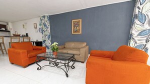 Una sala de estar con dos sofás naranjas, una mesa de centro de vidrio, un sofá color crema y una cortina estampada, además de un cuadro abstracto enmarcado en la pared.