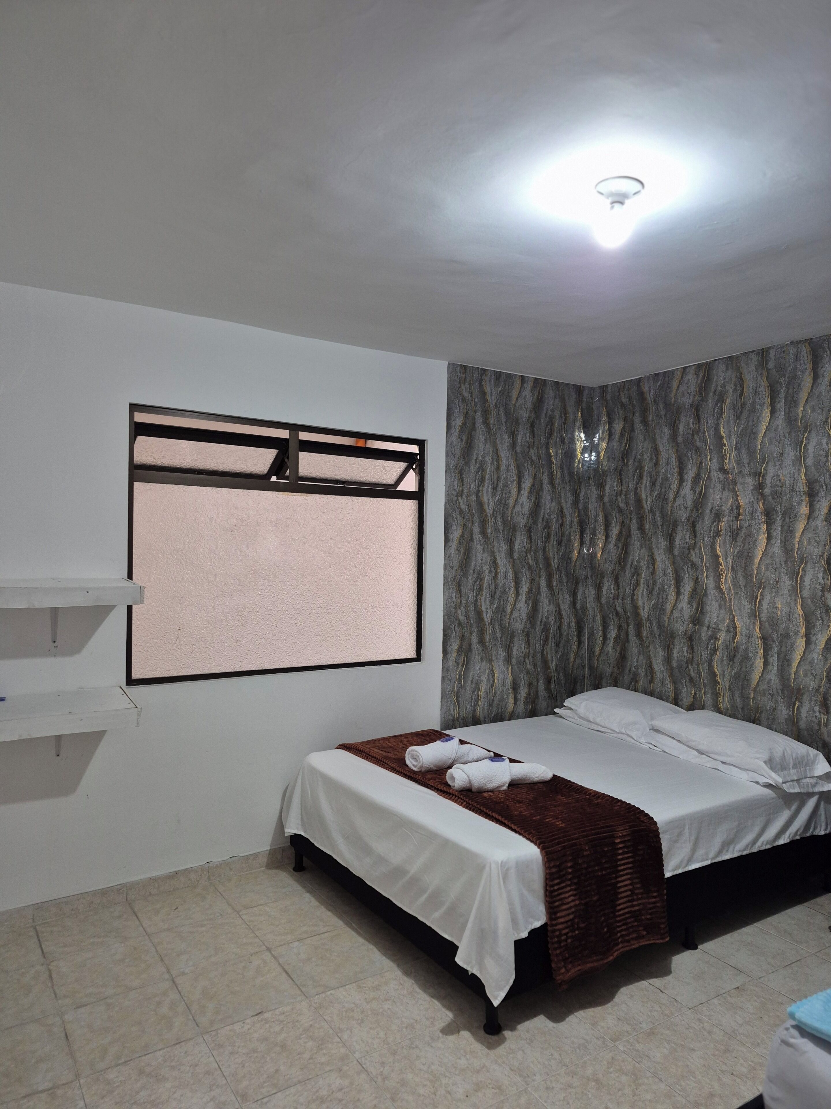 A bedroom with a bed, a window with blinds, a wall-mounted shelf, and a textured wall.