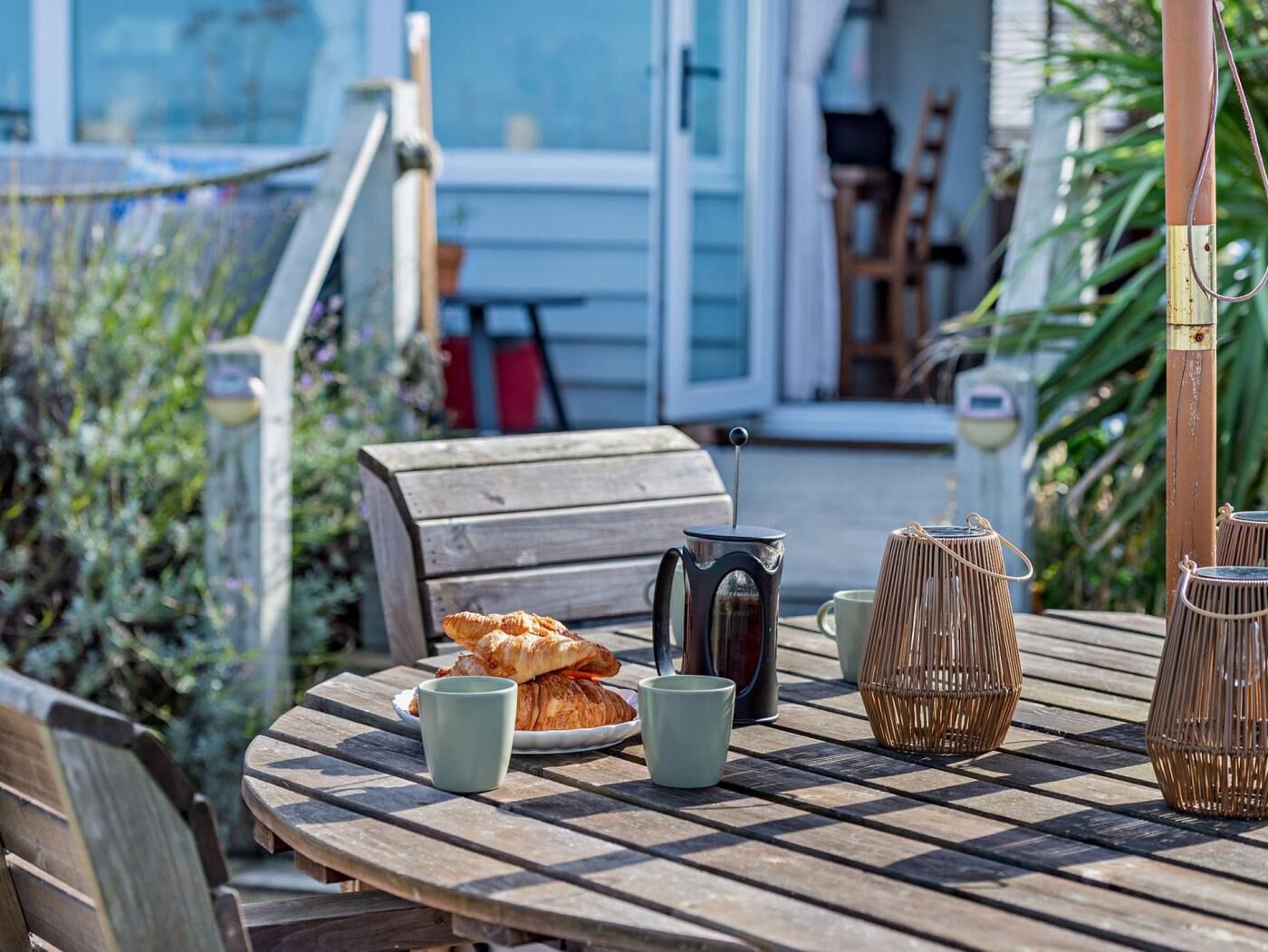 Une table en bois avec un croissant, une cafetière et deux tasses, disposée à l’extérieur, avec un banc et des vases en osier en arrière-plan.