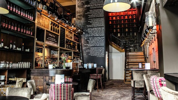 Um bar com prateleiras cheias de garrafas, um menu em lousa e móveis de vime.