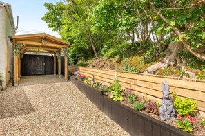 A covered outdoor area with a gravel path leading to a wooden structure, surrounded by a garden with various plants and a wooden fence. - 1 Bed in Stratton (oc-f35146) (Stratton)