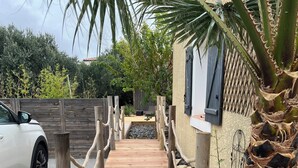 Une terrasse en bois mène à un bâtiment dont la façade présente un relief particulier, avec un palmier et une voiture garée à proximité.