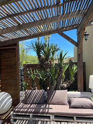 Een houten pergola met een bed en kussens, omringd door planten en een helderblauwe lucht.
