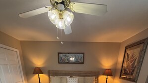 A bedroom with a wooden bed, a bench, two bedside lamps, a ceiling fan, and wall art. - D 32 Sundown Condominiums (Perdido Key)