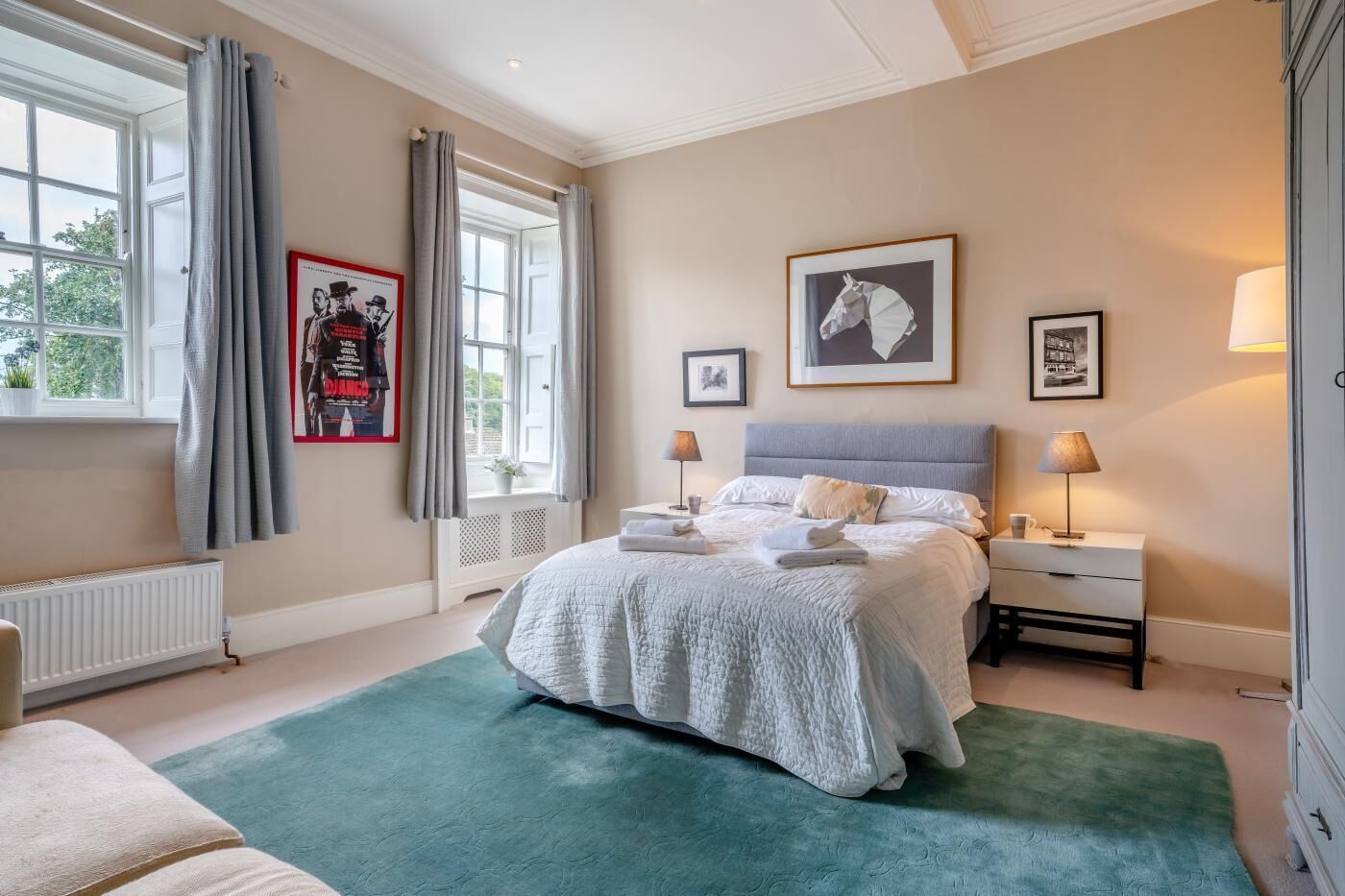 Une chambre à coucher comprenant un lit, deux tables de chevet, un radiateur, une fenêtre avec des rideaux et des œuvres d’art murales.