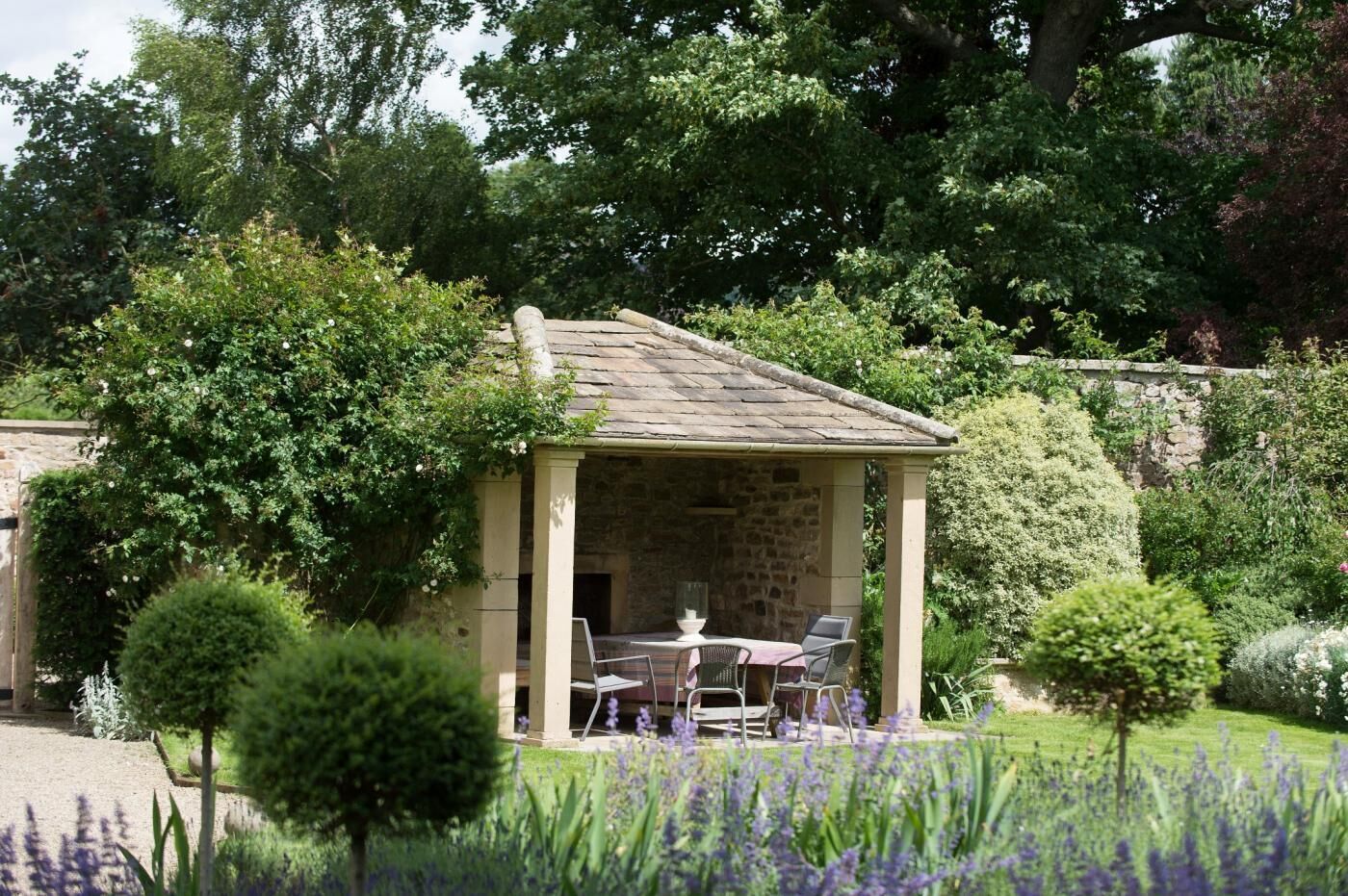 A stone-built gazebo with a table and chairs, surrounded by lush greenery and flowering plants.