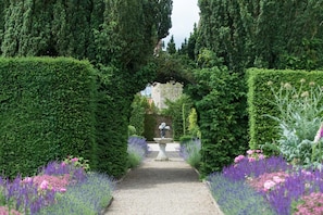 Ein Gartenweg mit einer Statue, umgeben von ordentlich geschnittenen Hecken und blühenden Pflanzen.