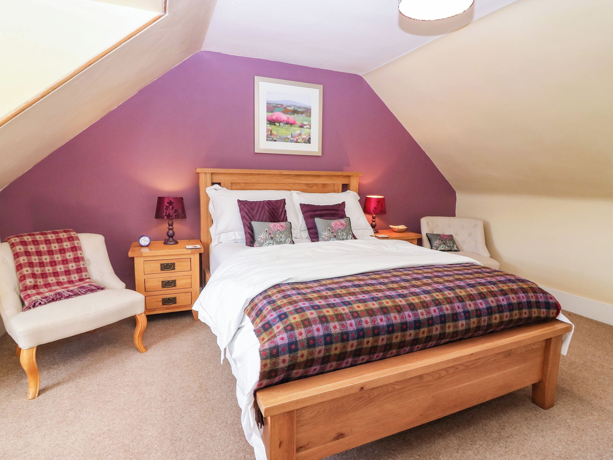 A bedroom with a sloped ceiling, a bed with a colorful blanket, two bedside tables with lamps, and a chair with a checkered blanket.