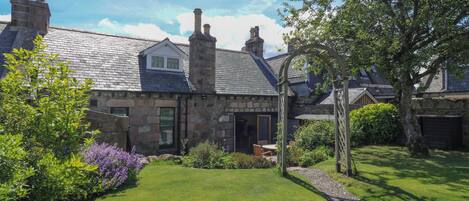 A stone house with a sloped roof, a garden with a lawn and shrubs, and a wooden pergola.