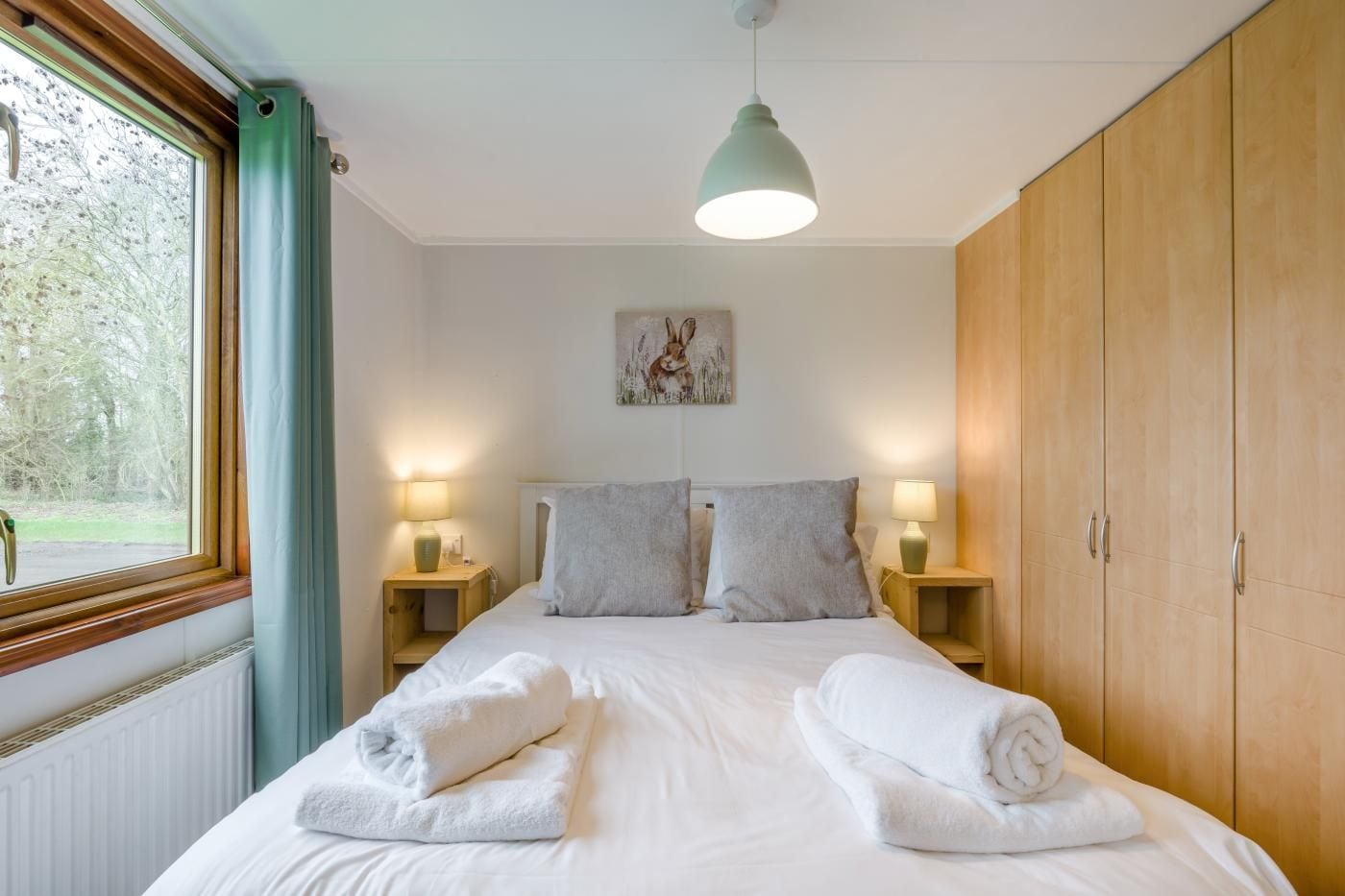 A bedroom with a bed, two bedside tables, a hanging light fixture, a wardrobe, and a window with curtains.