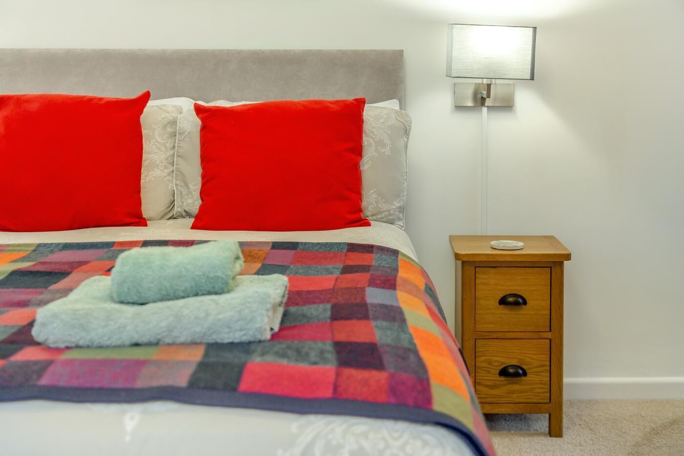 A neatly made bed with a colorful quilt, two red pillows, a bedside table with a lamp, and a small drawer.
